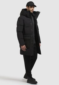 Sort dunjakke med hætte, lynlås og knaplukning samt store frontlommer. Båret over en grå sweater og parret med sorte bukser og støvler.