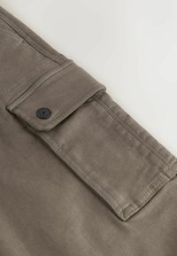 Tissu gris avec une texture lisse, comportant une poche à rabat fixée par un bouton noir. Détail de couture le long des bords.