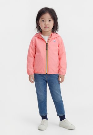 Malé dieťa stojí oblečené v korálovej bunde s kapucňou, modrých rifle, tmavých ponožkách a bielych teniskách na jednoduchom svetlom pozadí.