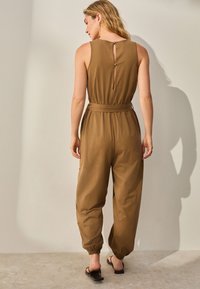 Pruun varrukateta kombinesoon, vöökohal vöö, tagaküljel avaus, elastsed varrukaotsad ja lõdvestunud lõige. Minimalistlik disain, sile tekstuur.