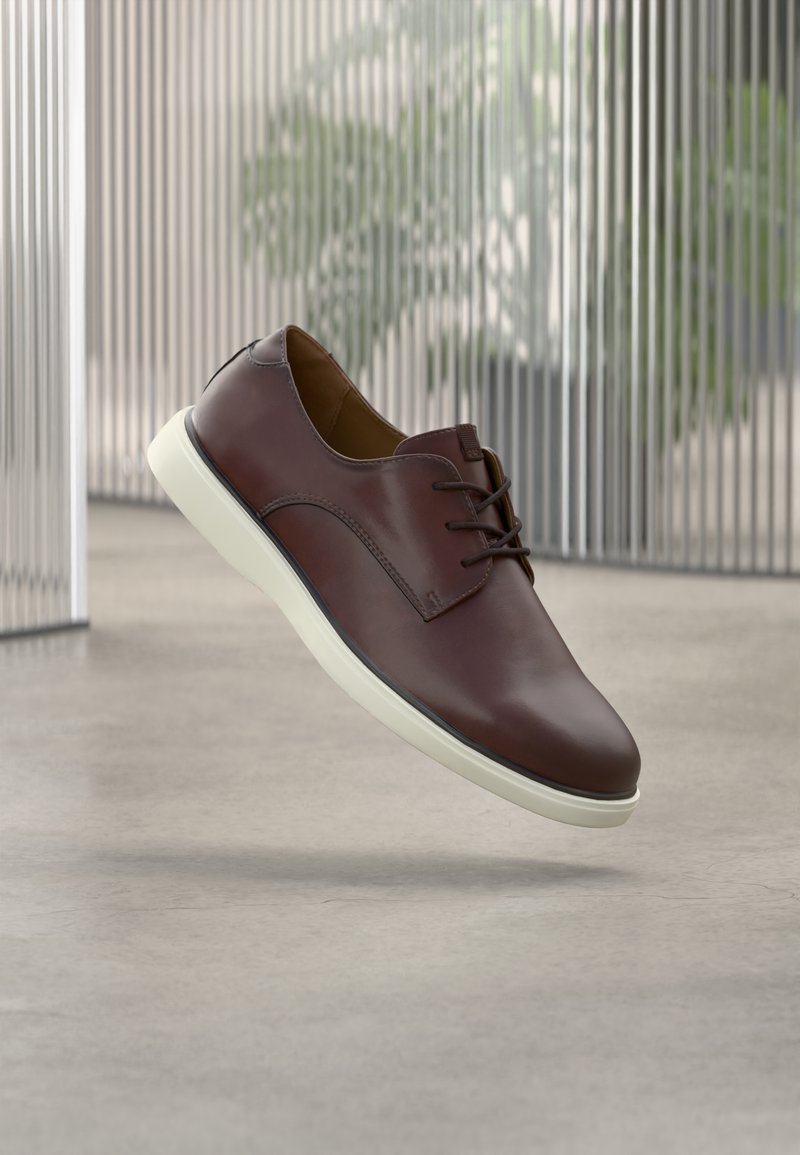 Chaussure à lacets en cuir marron avec semelle blanche suspendue au-dessus d'un sol en béton poli, avec un fond de lattes métalliques verticales et un arrière-plan flou de verdure.