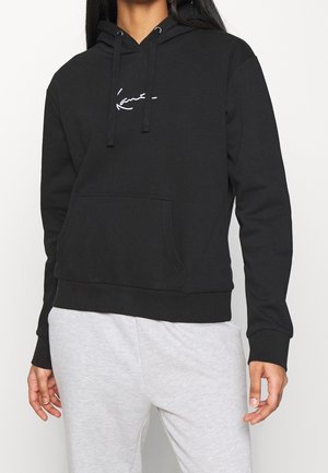 Personne portant un sweat à capuche noir avec une poche avant et un logo blanc en script, associé à un pantalon de jogging gris clair, visible du cou aux hanches.