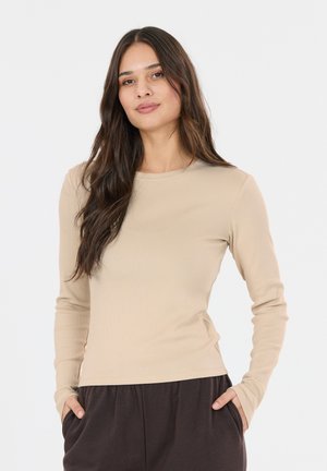 Ung kvinde med langt mørkt hår iført en beige langærmet trøje og mørke bukser, hænderne i lommerne, stående mod en hvid baggrund.