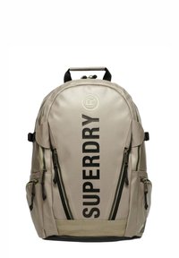Zaino beige con scritta nera "SUPERDRY", molteplici tasche con zip, materiale in tessuto, spallacci imbottiti e maniglia superiore per il trasporto.