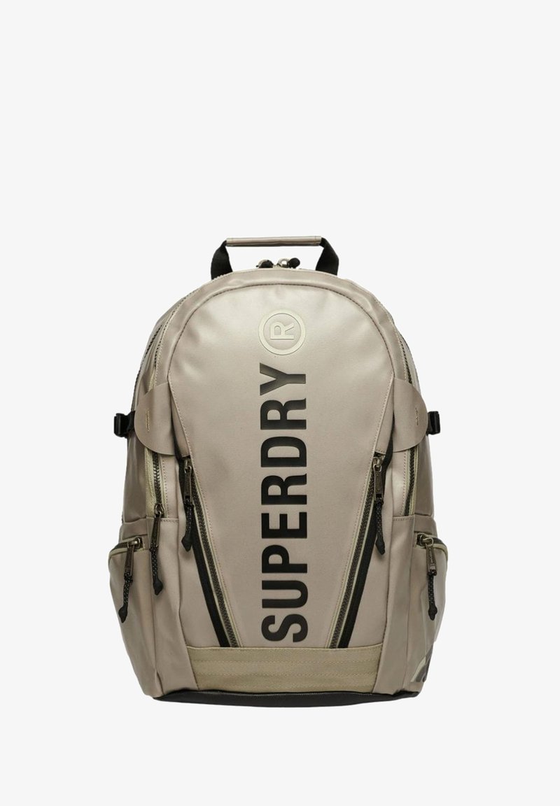 Zaino beige con scritta nera "SUPERDRY", molteplici tasche con zip, materiale in tessuto, spallacci imbottiti e maniglia superiore per il trasporto.