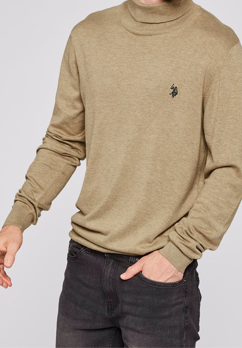 Homme portant un pull à col roulé beige avec un petit logo noir sur la poitrine, jean foncé, main gauche dans la poche, bras droit légèrement étendu.