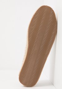 Vue de dessous d'une chaussure avec une semelle en caoutchouc naturel présentant un motif de bande de roulement ondulé, appuyée contre un mur blanc.