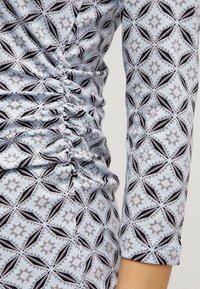 Långärmad klänning i ljusblått tyg med ett geometriskt mönster i svart och beige, med rynkdetaljer längs sid sömmarna.