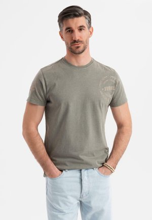 Olivengrünes Baumwoll-T-Shirt mit kurzen Ärmeln, rundem Ausschnitt und Logodetail in hellbeige. Kombiniert mit hellblauen Jeans.