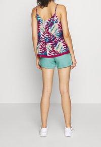 Halenka bez rukávů s peplum střihem v bílé barvě s barevným květinovým vzorem, doplněná o světle zelené šortky. Modelka má na sobě bílé tenisky.
