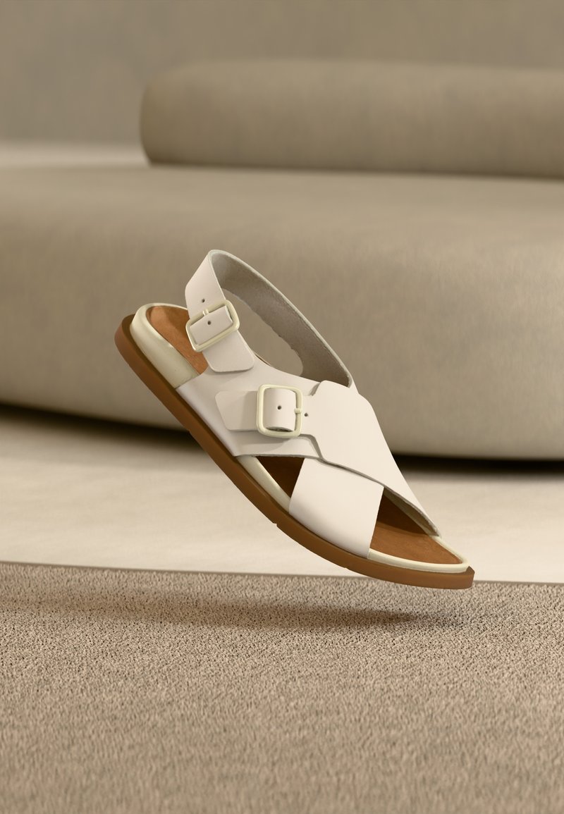 Λευκό δερμάτινο σανδάλι με δύο ρυθμιζόμενες αγκράφες, ανοιχτό σχέδιο στα δάχτυλα και καφέ λαστιχένια σόλα, τοποθετημένο πάνω σε υφασμένη μπεζ επιφάνεια.