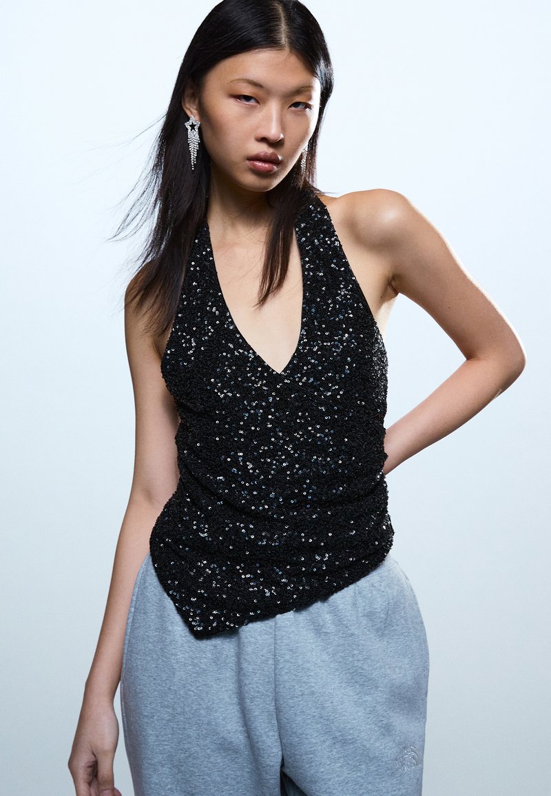 Νεαρή γυναίκα με μακριά σκούρα μαλλιά φοράει μαύρο αμάνικο τοπ με παγιέτες, γκρι φόρμα και κρεμαστά σκουλαρίκια σε σχήμα αστεριού, μπροστά σε ανοιχτό φόντο.