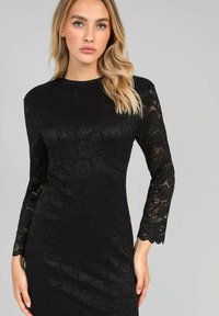 Robe en dentelle noire à manches longues, ornée d'un motif floral et de bords festonnés. La coupe ajustée met en valeur la silhouette.