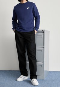 Sweatshirt bleu marine avec le logo Nike blanc, associé à un pantalon noir et des sneakers blanches. Se tenant à côté d'un meuble de rangement gris.