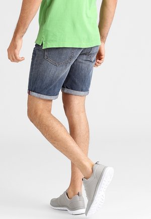 Jeansowe szorty w ciemnym niebieskim kolorze z podwiniętym dołem, zestawione z zieloną koszulą i jasnoszarymi trampkami, prezentujące swobodny letni styl.