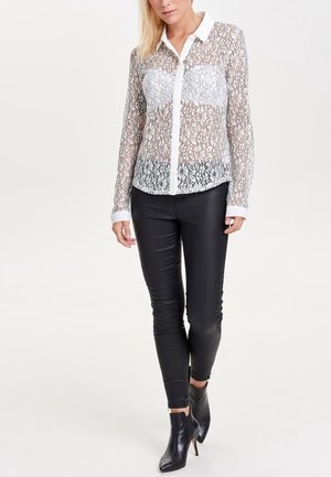 Blusa bianca trasparente di pizzo con motivi floreali, maniche lunghe e colletto classico, abbinata a pantaloni di pelle nera e stivaletti.