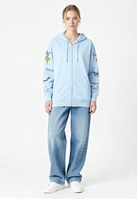 Sweat à capuche zippé bleu clair avec différentes impressions colorées sur les manches, associé à un jean délavé à coupe ample et des baskets blanches.