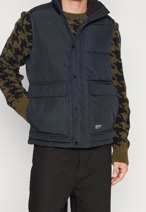 Man draagt een zwart doorgestikt vest over een groene en zwarte camouflage trui met een zwarte broek, hand rust op de heup.