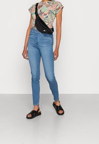 Conjunto informal que incluye una blusa de manga corta con un patrón colorido, unos jeans ajustados de talle alto en azul y sandalias negras de tiras cruzadas.