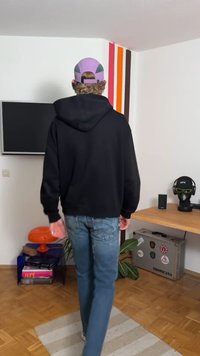 Černá mikina s kapucí, modré džíny a levandulová čepice. Pokoj obsahuje televizi, dřevěný stůl s dekoracemi a květináč s rostlinou.