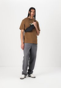 Brun bomulds t-shirt med grafisk design, grå denim jeans og sort talje taske. Modellen har hvid og grå sneakers på og står foran en ensfarvet baggrund.