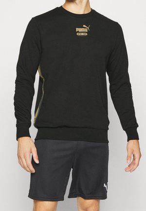 Man draagt een zwarte Puma King longsleeve trui en zwarte Puma shorts tegen een effen achtergrond.