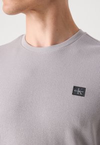 Pelēks ribots T-krekls ar apaļu kaklu, kas akcentēts ar Calvin Klein logo uz kreisās puses. Audums šķiet mīksts ar teksturētu apdari.