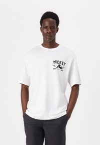 Hvid bomuld t-shirt med rund hals, korte ærmer, prydet med et sort grafiktryk, der siger "MICKEY" samt en silhuet af Mickey Mouse.