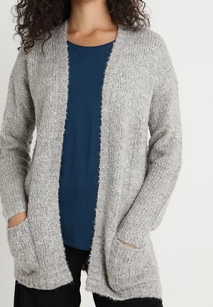 Frau trägt einen weichen, hellgrauen Cardigan mit offenem Vorderteil über einem dunkelblauen Hemd, die Hände in den Taschen, vor einem schlichten Hintergrund stehend.