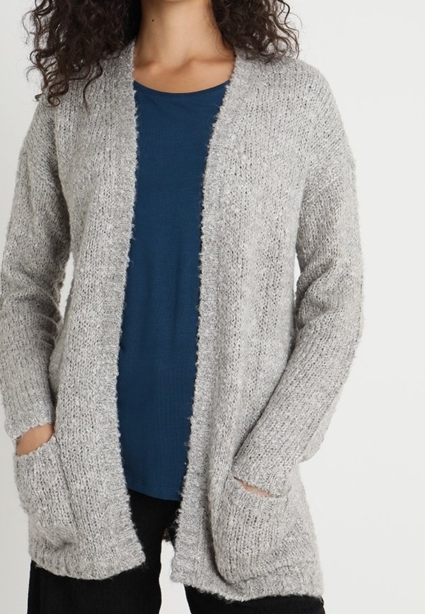 Femme portant un cardigan doux, gris clair, ouvert devant, par-dessus une chemise bleu foncé, les mains dans les poches, debout devant un fond uni.