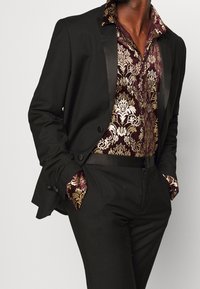 Černý oblek s lesklým saténovým límcem, doplněný bordó košilí s vzorem a zlatými detaily. Slim fit střih s propracovanými manžetami.