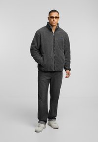 Gray fleece jacket with high collar and front zipper, featuring side pockets and a patch on the chest. Paired with dark jeans and white shoes.