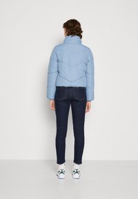 Světle modrá péřová bunda s vysokým límcem, šikmým prošíváním a elastickými manžetami, kombinovaná s tmavými džíny a bílými teniskami.