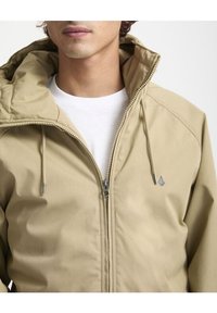 Giacca beige con zip e collo alto, dotata di coulisse, tessuto liscio e piccolo logo sull'area sinistra del petto.