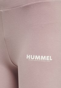 Różowe legginsy wykonane z miękkiego, elastycznego materiału o gładkiej fakturze. Posiadają białe logo "HUMMEL" z napisem "SPORTSWEAR SINCE 1923" w pogrubionej czcionce.