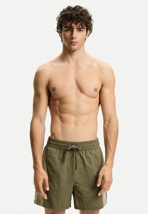 Men's olive green swim shorts with elastic waistband and drawstring. Features lightweight textured fabric and side color-block accents.