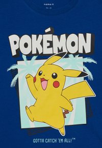 Blå bomull t-skjorte med grafikk av Pikachu, med teksten "POKÉMON" og "MÅ FANGE DEM ALLE!" i fete hvite bokstaver.