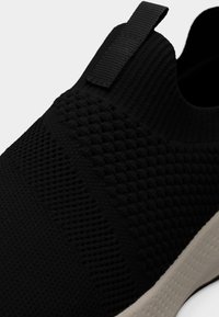 Κοντινό πλάνο μαύρου πλεκτού αθλητικού παπουτσιού χωρίς κορδόνια, με υφή στην επάνω επιφάνεια και γκρι ανοιχτό λαστιχένια σόλα, με θηλιά στη φτέρνα για εύκολη φορεσιά.