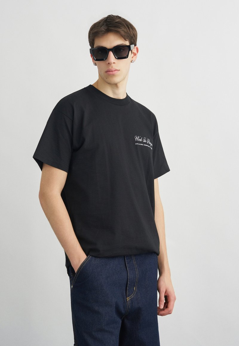 Νεαρός άνδρας που φοράει μαύρα γυαλιά ηλίου, μαύρο μπλουζάκι με γραφικά και σκούρο μπλε τζιν, στέκεται με το ένα χέρι στην τσέπη μπροστά σε μια απλή φόντο.