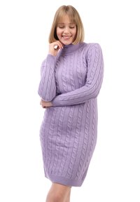 Frau in einem lilafarbenen, strukturierten Rollkragenpulloverkleid, die lächelt, während sie mit einer Hand ihr Kinn berührt und den anderen Arm verschränkt.
