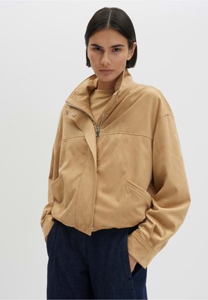 Frau mit kurzen dunklen Haaren trägt eine beige Jacke mit Reißverschluss und hohem Kragen sowie dunkelblaue Hose, Hände in den Taschen, vor einem schlichten hellen Hintergrund.