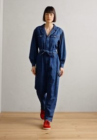 Denim jumpsuit featuring long sleeves, two chest pockets, a belted waist, and wide legs. Worn with red shoes on a wooden floor.