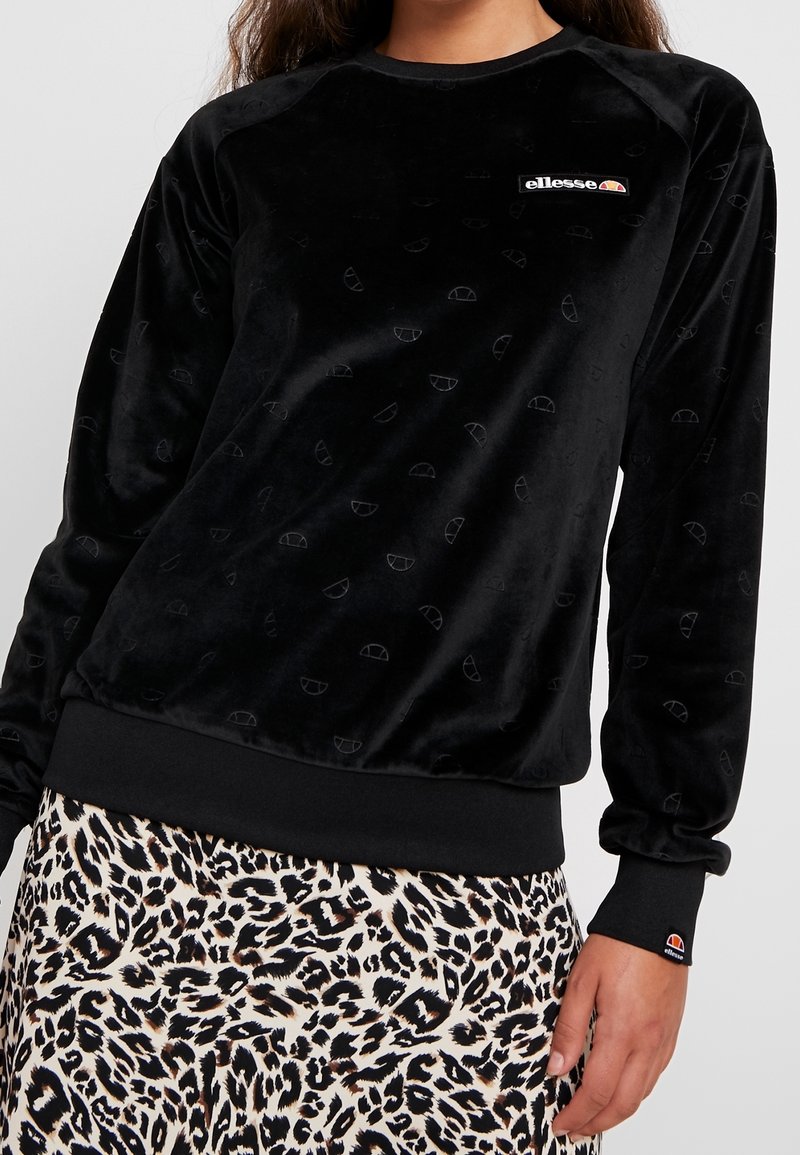 Sweat-shirt noir velouté manches longues avec des logos subtils et un patch blanc avec le logo ellesse, associé à une jupe à imprimé léopard.