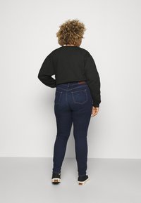 Schwarzes Langarmshirt, dunkelblaue enge Jeans mit Hintertaschen und Steppdetails, kombiniert mit schwarzen Schuhen. Einfaches und figurbetontes Design.