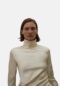 Kremna turtleneck majica iz gladkega materiala, z dolgimi rokavi in prilegajočim se krojem. Preprosta, minimalistična zasnova brez vidnih vzorcev.
