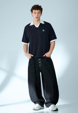 Mlad moški stoji z rokami v žepih, nosi ohlapne črne kavbojke, mornarsko modro kratko majico s belim ovratnikom in bele superge, nevtralno ozadje.