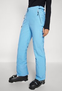 Pantaloni de schi de culoare albastru deschis, din țesătură rezistentă la apă, cu multiple buzunare cu fermoar și talie ajustabilă. Purtați cu bocanci de schi negri.