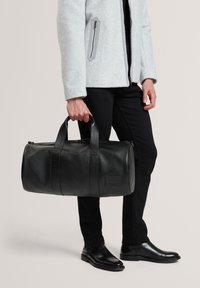 Sac de voyage en cuir noir à fini texturé, avec double poignée et fermeture à glissière. Porté avec une veste grise et un pantalon noir, associé à des bottes polies.