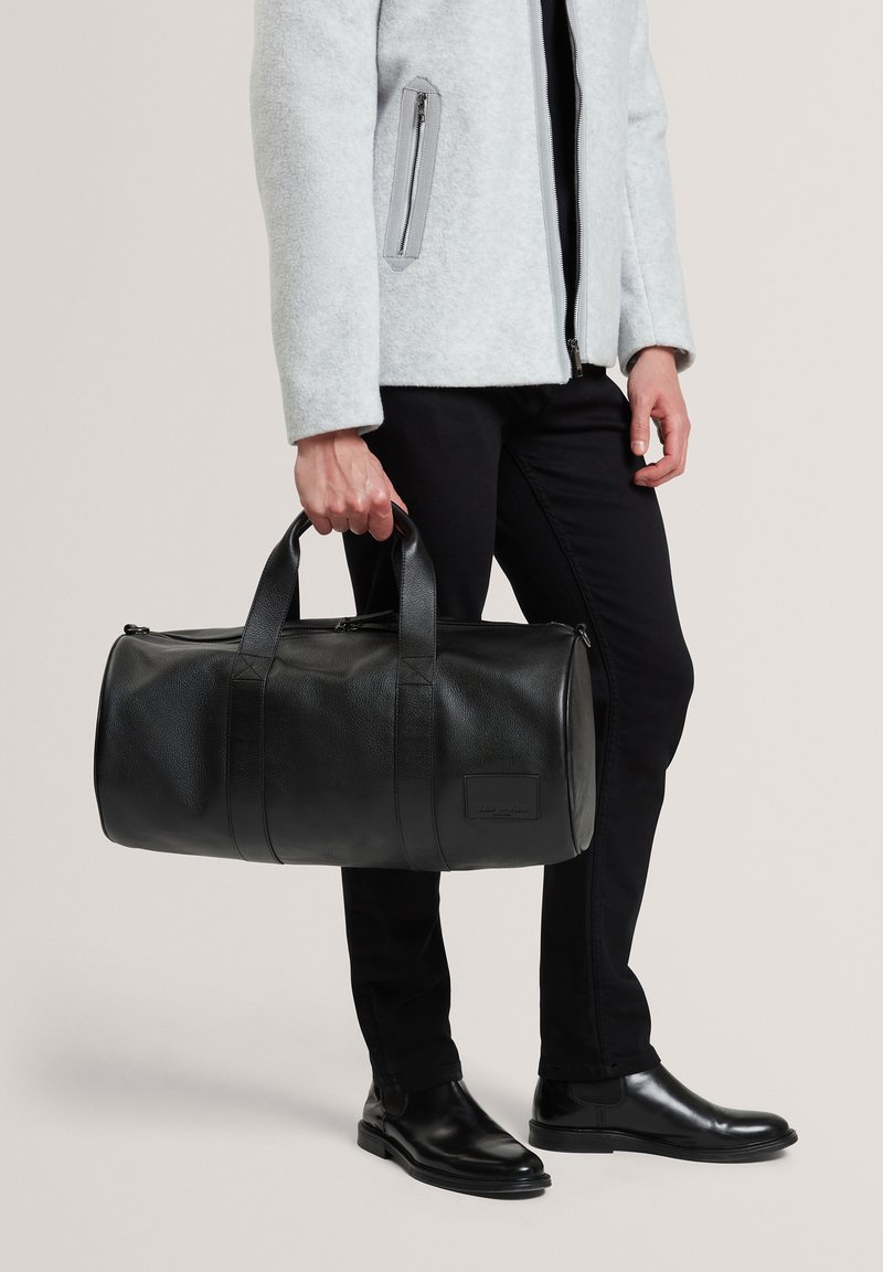Sac de voyage en cuir noir à fini texturé, avec double poignée et fermeture à glissière. Porté avec une veste grise et un pantalon noir, associé à des bottes polies.