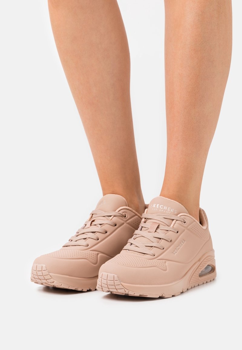 Jambes portant des baskets à lacets Skechers rose clair avec semelles épaisses et coussin d’air visible, debout sur fond blanc.